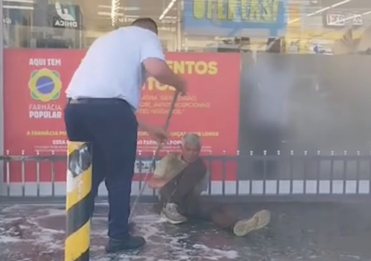 Funcionário limpa calçada com homem em situação de rua no local